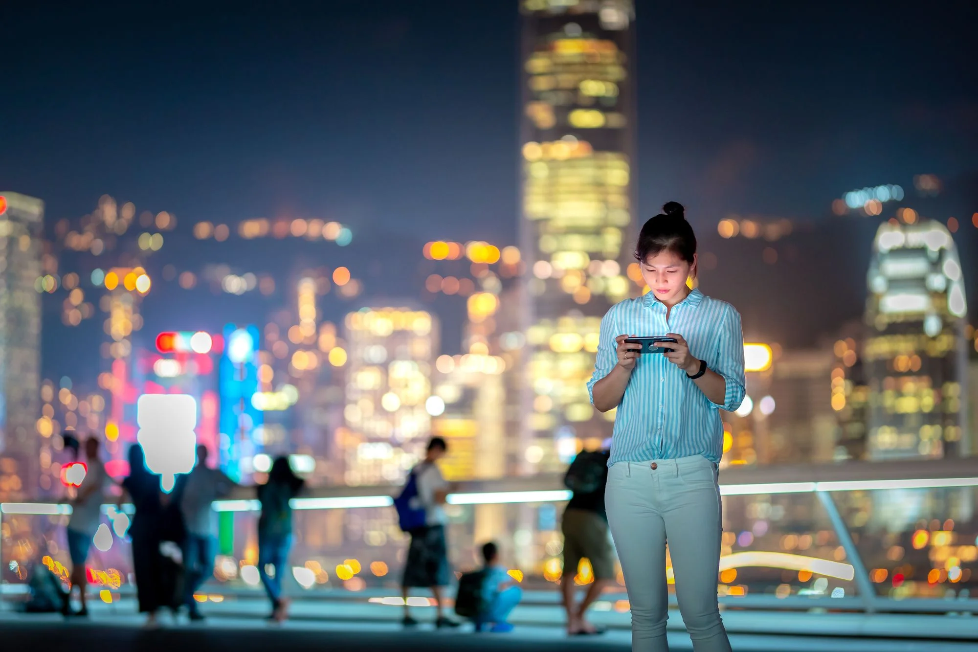 Woman using smartphone at night with Hong Kong's Victoria Harbor in background.